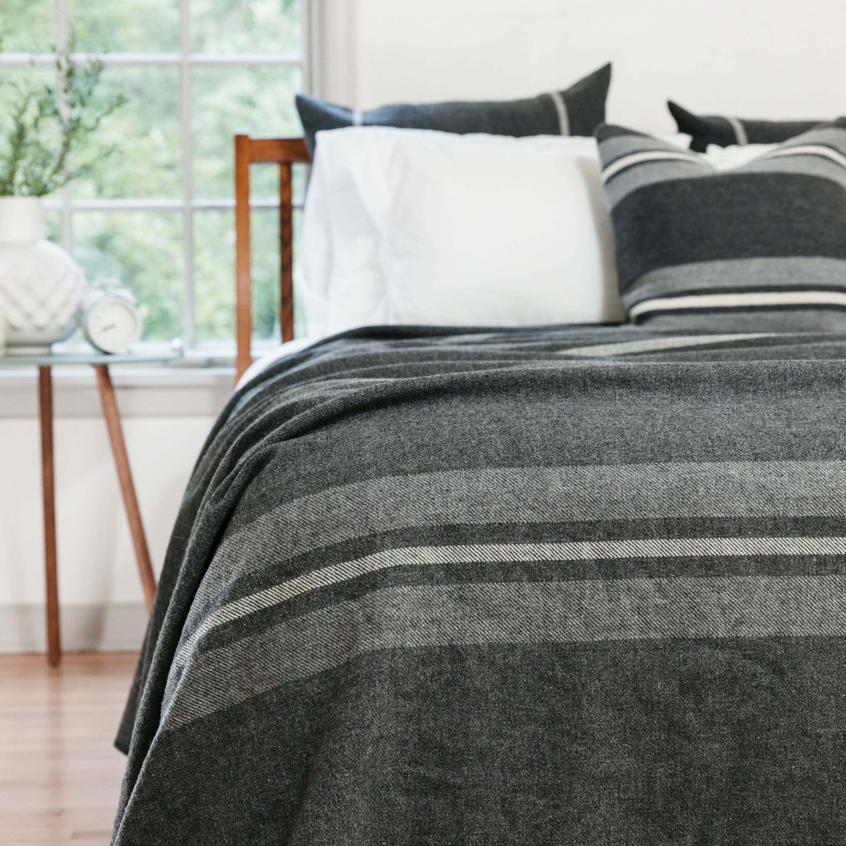 Cabin Charcoal middleweight wool blanket with heather gray and natural stripes on a bed, styled with grey striped pillows next to a glass table.