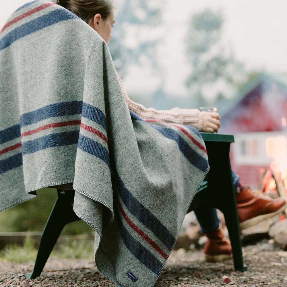 Cabin throw wool blanket in heather gray with navy and red stripes wrapped around a person on an Adirondack chair by an outdoor campfire with a red cabin in the background.