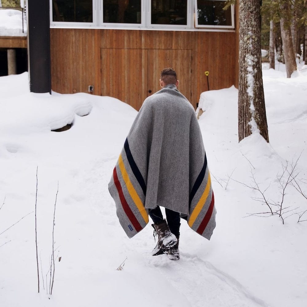 Frontier Gray wool throw blanket with navy, gold, and red stripes wrapped around a man walking through deep snow toward a wooden cabin in the woods.