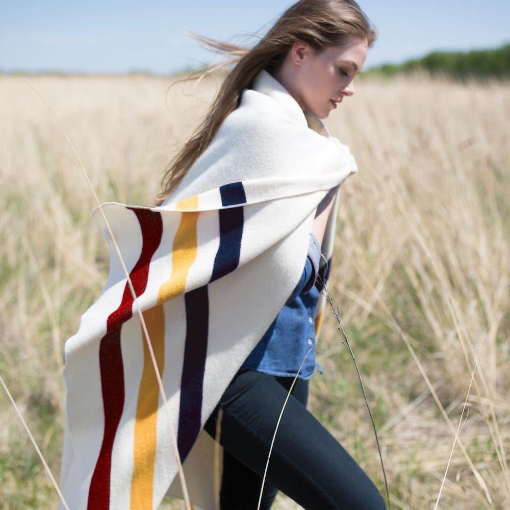 Frontier Natural wool throw blanket with navy, gold, and red stripes wrapped around a woman walking through a windy, yellow wheat field.