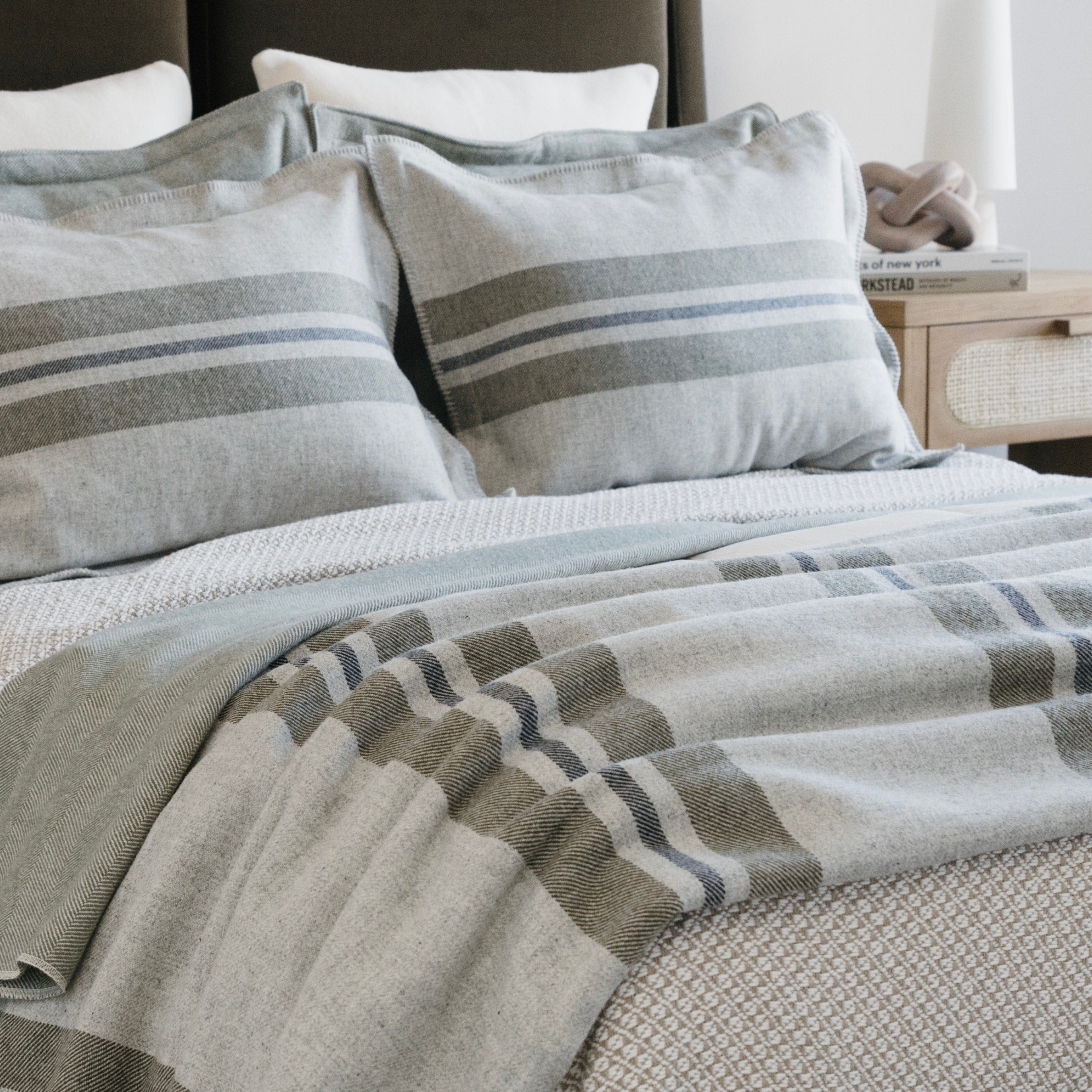 Cabin middleweight wool blanket in smoke with olive and navy stripes on a bed, with matching striped pillows and a nightstand with books and a lamp.