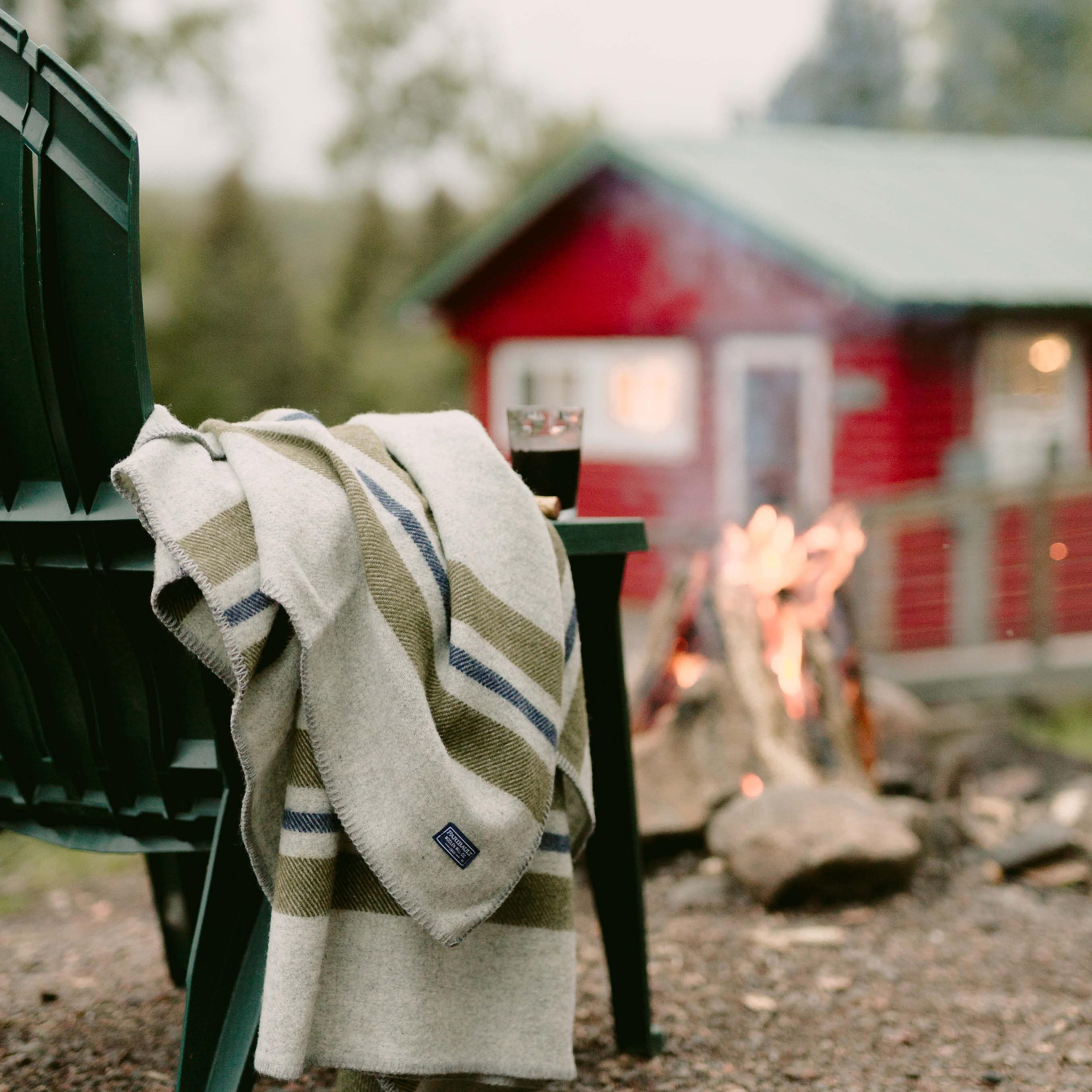 Cabin throw wool blanket in smoke with olive and navy stripes on an Adirondack chair by an outdoor campfire with a red cabin in the background.