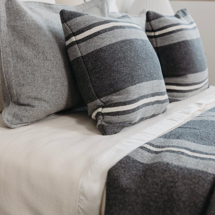 Pure and Simple Satin Bone White middleweight wool blanket styled as a layering blanket under a charcoal blanket on a bed with gray striped pillows.