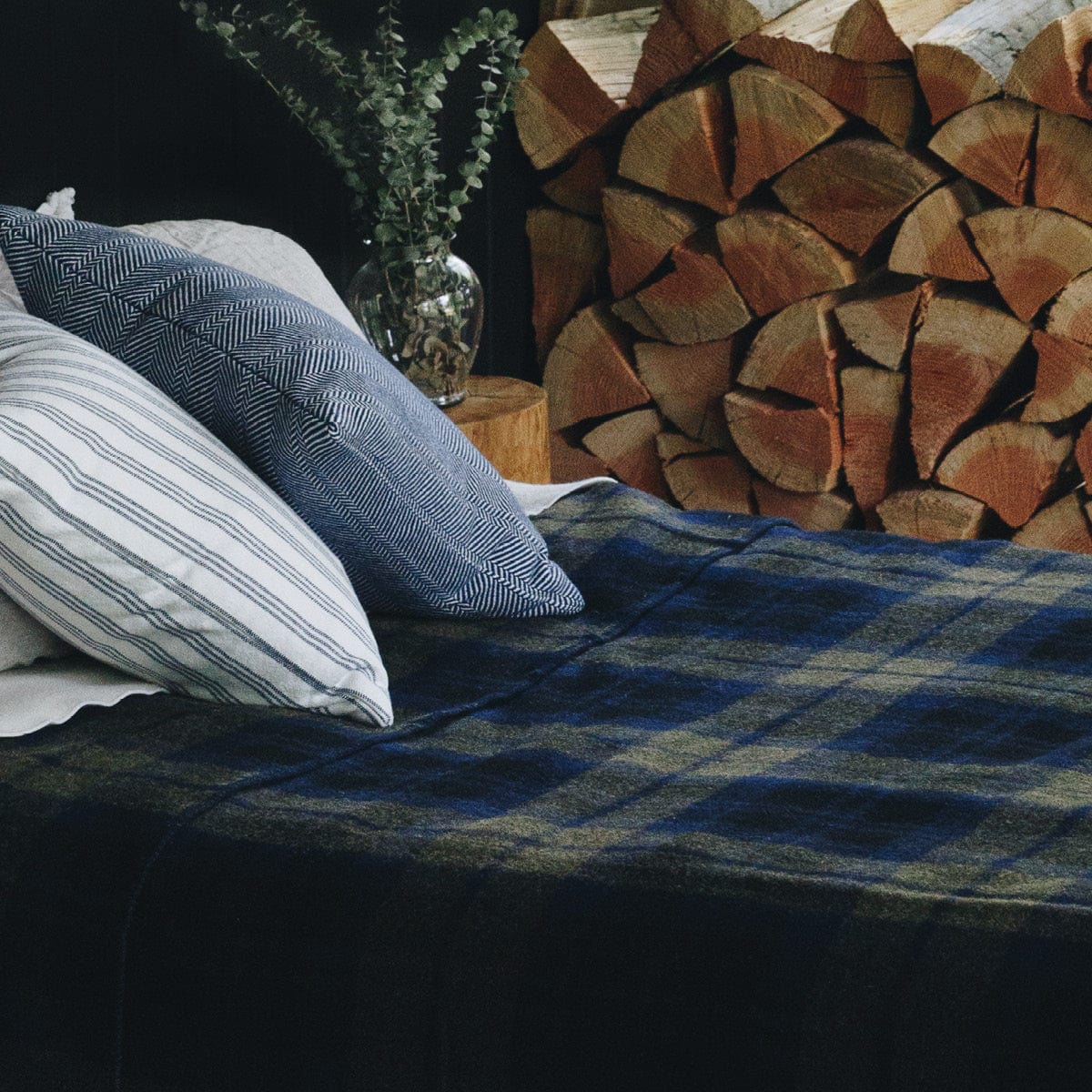Shadow Plaid green middleweight wool blanket on a bed, styled with striped pillows and a plant vase on a log nightstand next to stacked firewood.