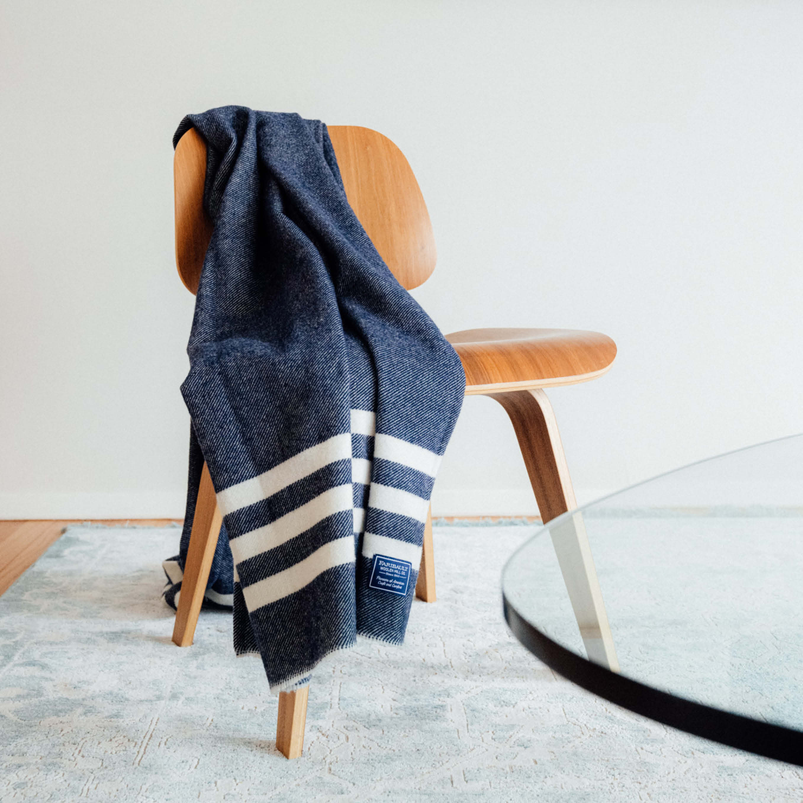 Trapper Navy lightweight wool blanket throw with natural stripes draped on a wooden mid-century chair over a light blue rug next to a glass table.